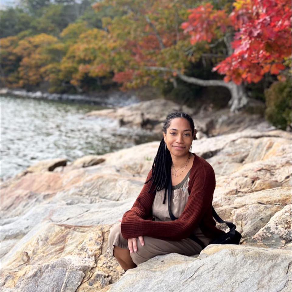 Photo of Malika Davis on rock by riverside with fall foliage in the background | Black Owned Maine Portland