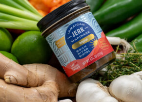 Photo of a jar of Jamaican jerk marinade from Back River Blends - background is a bit blurred but the jar is on top of ginger root, garlic bulbs, thyme springs, peppers, and limes | Black Owned Maine Portland