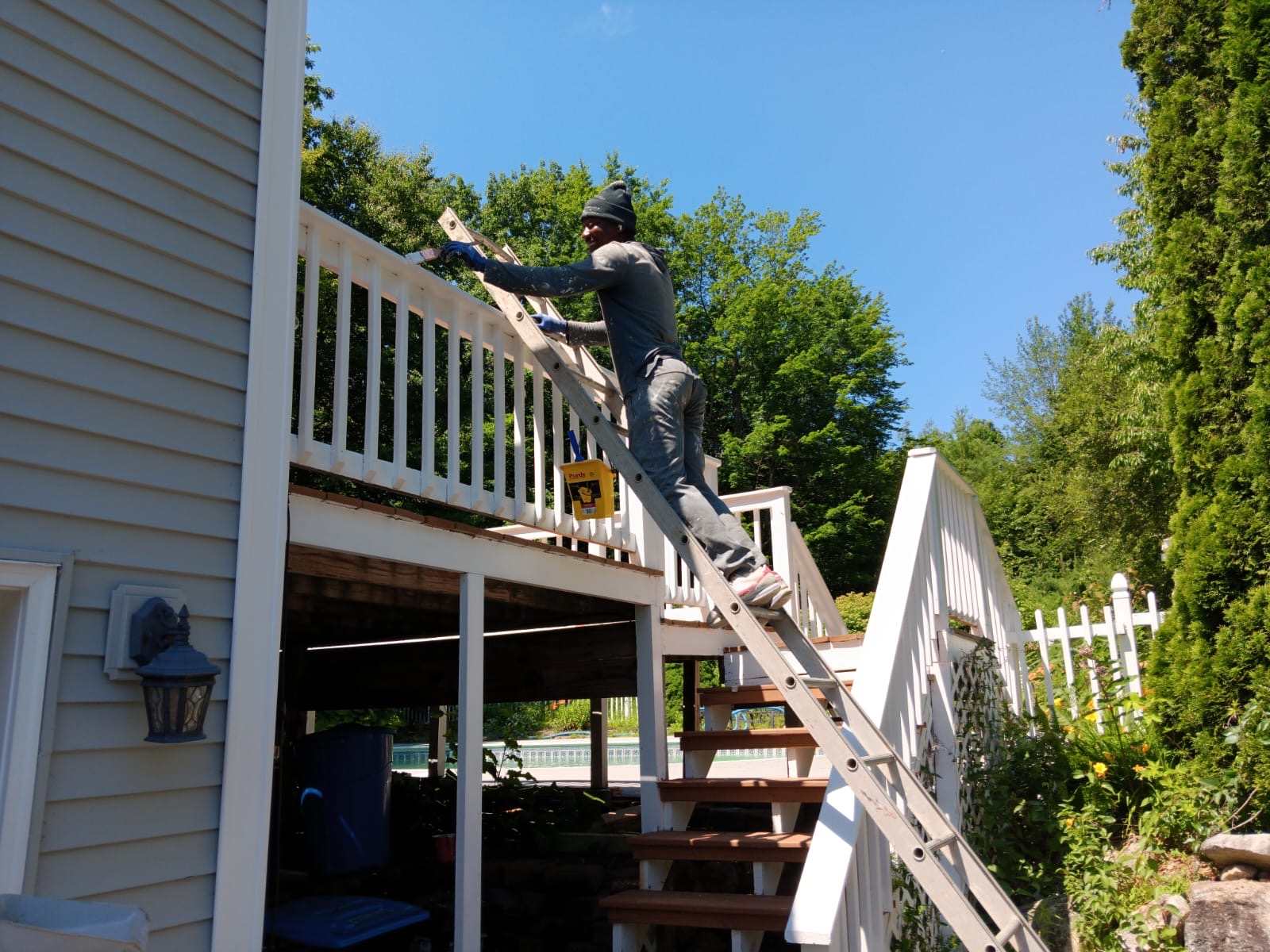 Photo of a man on a ladder painting a two-story deck of a grey house with greenery surrounding him | Black Owned Maine Portland