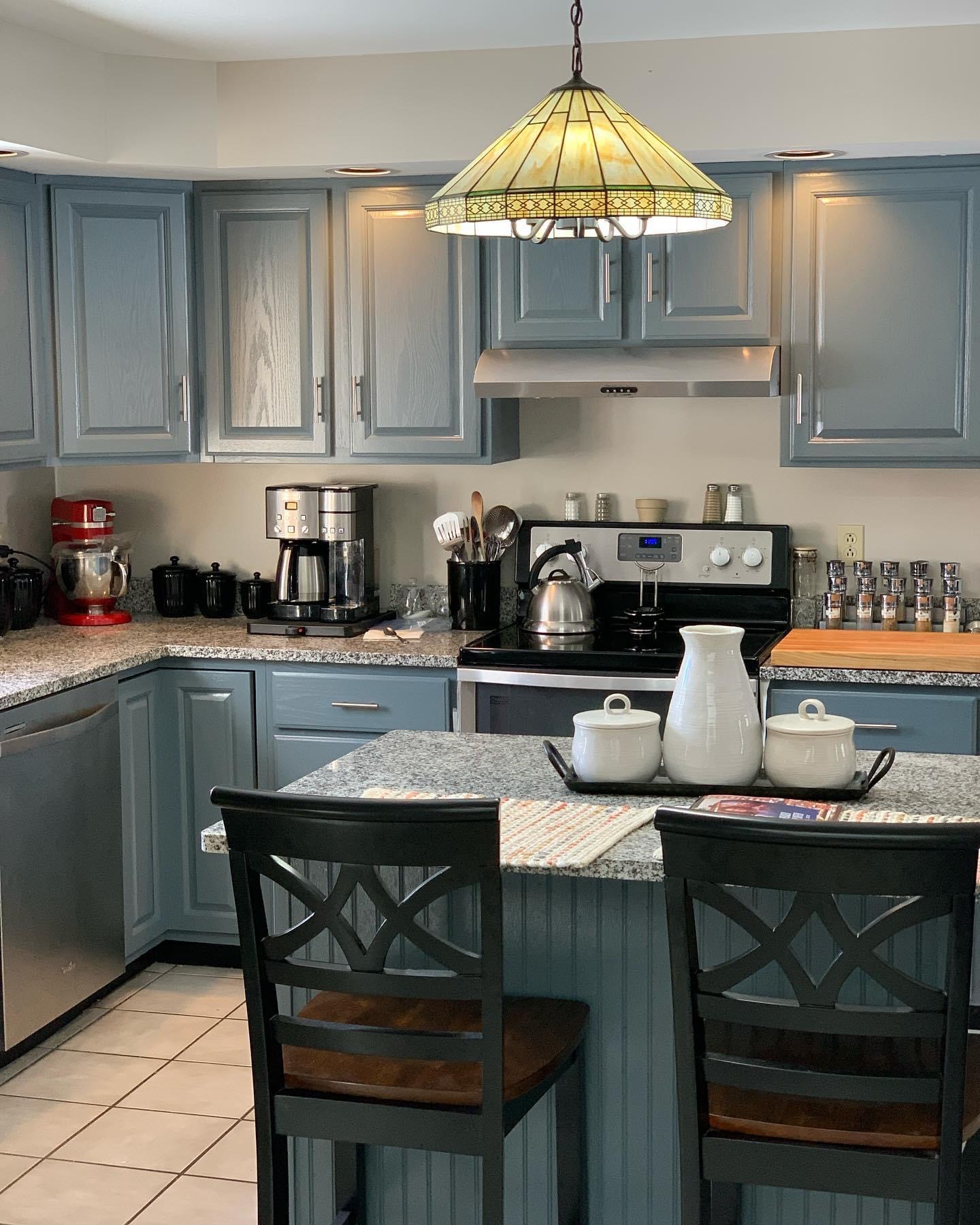 Image of kitchen with blue gray cabinets and stained glass light over a bar with decorative items on it and two barstool chairs with black backs and brown seats. | Black Owned Maine